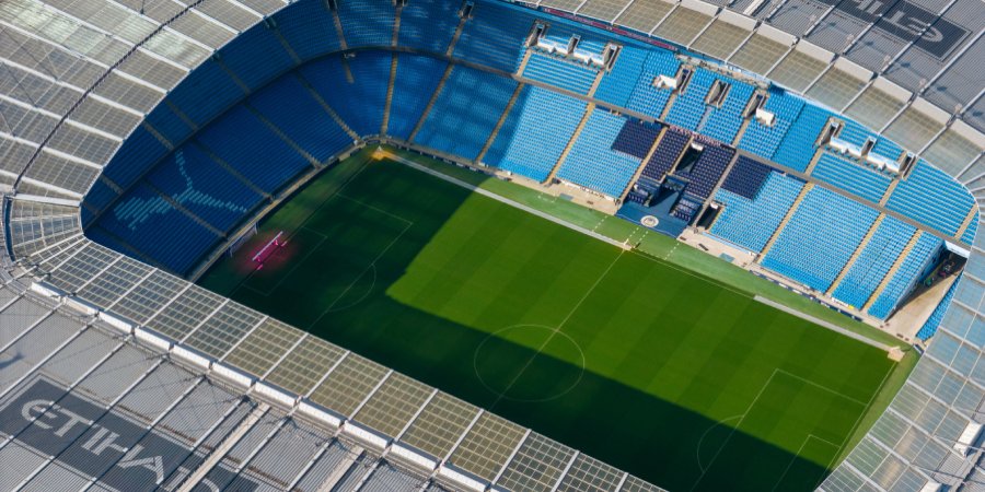 An aerial view of the empty Etihad Stadium showing the green pitch and blue seating under bright sunlight.